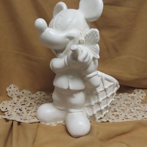 May include: A white ceramic figurine of Mickey Mouse holding a butterfly. The figure is wearing a suit and has a basket on its back. The figurine is standing on a decorative lace doily.