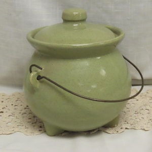 May include: A light green ceramic pot with a lid and a metal handle. The pot has three small feet and a rounded body. The lid has a small knob on top. The pot is sitting on a cream-colored lace doily.