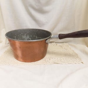 以下が含まれることがあります： 濃い木製のハンドルと金属製のブラケットが付いた銅製のソースパン。鍋の内側には使用感が見られます。鍋はクリーム色のレースのドイリーの上に置かれています。