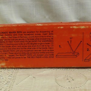 May include: An orange box with text and diagrams for sharpening knives. The text reads "COLORADO BEAVER TEETH" and instructions for use. Diagrams show the sharpening process. The box is on a white lace surface.