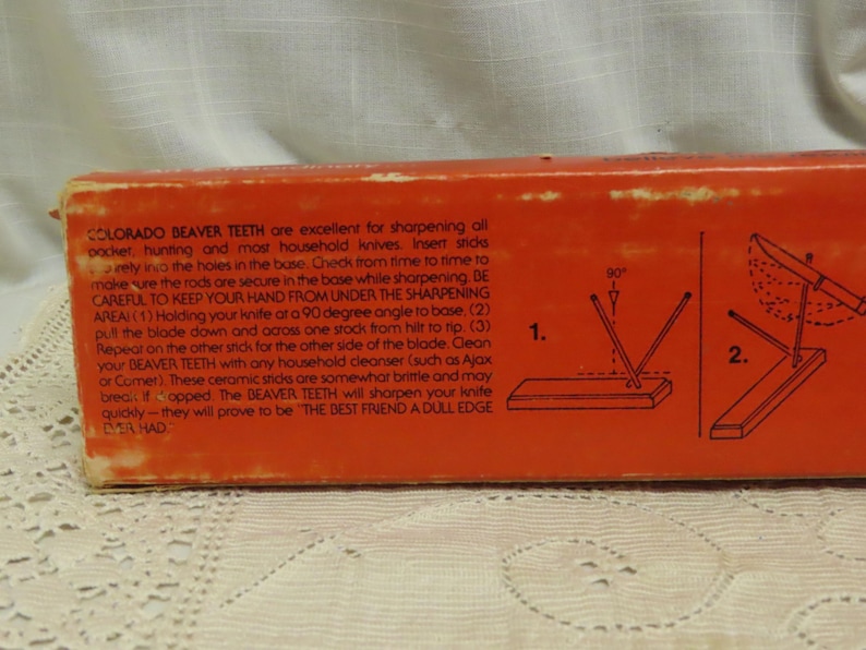 May include: An orange box with text and diagrams for sharpening knives. The text reads "COLORADO BEAVER TEETH" and instructions for use. Diagrams show the sharpening process. The box is on a white lace surface.