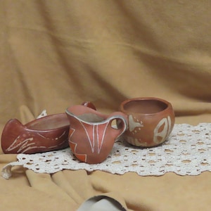 Könnte beinhalten: Drei handgefertigte, rötlich-braune Keramikstücke mit weiß bemalten Designs. Die Artikel umfassen eine kleine bootsförmige Schale, einen Henkelbecher und eine runde Schüssel. Sie werden auf einer weißen Spitzendecke vor einem beigefarbenen Hintergrund präsentiert.