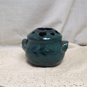 May include: A teal ceramic flower frog with a rounded body and two handles. The top has heart-shaped holes. Dark blue leaf designs are painted on the frog. A decorative item for floral arrangements.