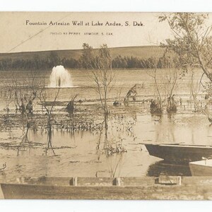Puede incluir: Una postal en blanco y negro que muestra un pozo artesiano de fuente en el lago Andes, Dakota del Sur. El pozo está en el medio de un lago rodeado de árboles y arbustos. Hay dos botes en primer plano.