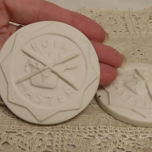 Op de afbeelding: Twee witte Boil Master onderzetters van keramiek. Elke onderzetter is rond met een opstaande rand en heeft de woorden "BOIL MASTER" en gekruiste breinaalden. De onderzetters liggen op een kanten oppervlak.