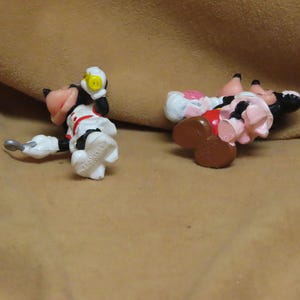 Puede incluir: Dos peque&ntilde;as y coloridas figuras de personajes de dibujos animados. Una est&aacute; vestida con un atuendo blanco con detalles rojos y un casco amarillo. La otra figura lleva un vestido rosa y un sombrero blanco. Ambas figuras est&aacute;n tumbadas sobre una superficie marr&oacute;n.