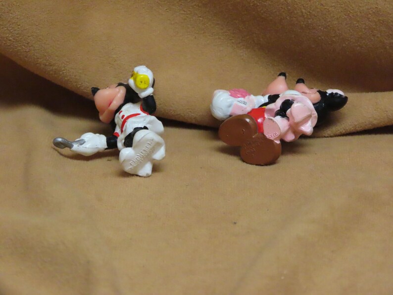 Puede incluir: Dos peque&ntilde;as y coloridas figuras de personajes de dibujos animados. Una est&aacute; vestida con un atuendo blanco con detalles rojos y un casco amarillo. La otra figura lleva un vestido rosa y un sombrero blanco. Ambas figuras est&aacute;n tumbadas sobre una superficie marr&oacute;n.