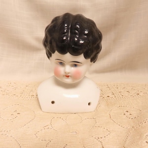 May include: A vintage doll head with black molded hair, blue eyes, and rosy cheeks. The porcelain head is white with a smooth finish and a small neck. The doll head has a classic, antique appearance, and is set against a cream-colored background.