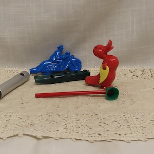 May include: A collection of vintage toy whistles and noisemakers. Includes a blue motorcycle, a red bird with yellow accents, a green log-shaped whistle, a silver whistle, and a red whistle with a green end.