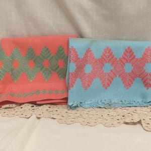 May include: Two rectangular fabric textiles featuring a repeating geometric diamond and leaf pattern. One textile is coral with mint green embroidery, while the other is light blue with coral embroidery. The blue textile has a fringed bottom edge.