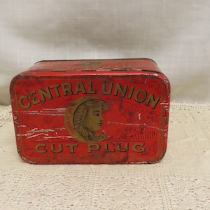 Puede incluir: Una lata rectangular roja vintage con las palabras "CENTRAL UNION CUT PLUG" en letras doradas. Una imagen circular del perfil de una mujer está en el centro. La lata tiene una apariencia ligeramente desgastada, lo que sugiere edad y uso.