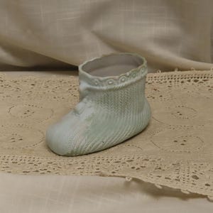 Könnte beinhalten: Hellblaue Baby-Keramikstiefel mit strukturiertem Strickmuster. Der Stiefel hat einen gewellten Rand und liegt auf einer cremefarbenen Spitzendecke.