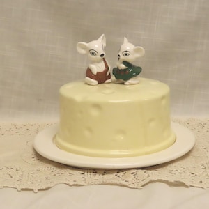 May include: A ceramic cheese dish with a yellow, cheese-like dome and a white plate base. Two small, white mouse figurines with brown and green outfits sit atop the dome. The dish is on a lace doily.
