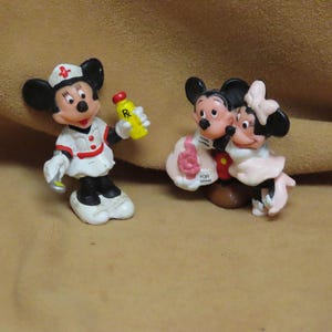 Puede incluir: Dos peque&ntilde;as figuras de Disney. Una es Mickey Mouse vestido de enfermera, sosteniendo una botella amarilla con una tapa roja. La otra figura es Mickey y Minnie Mouse abraz&aacute;ndose. Las figuras est&aacute;n sobre una superficie beige.