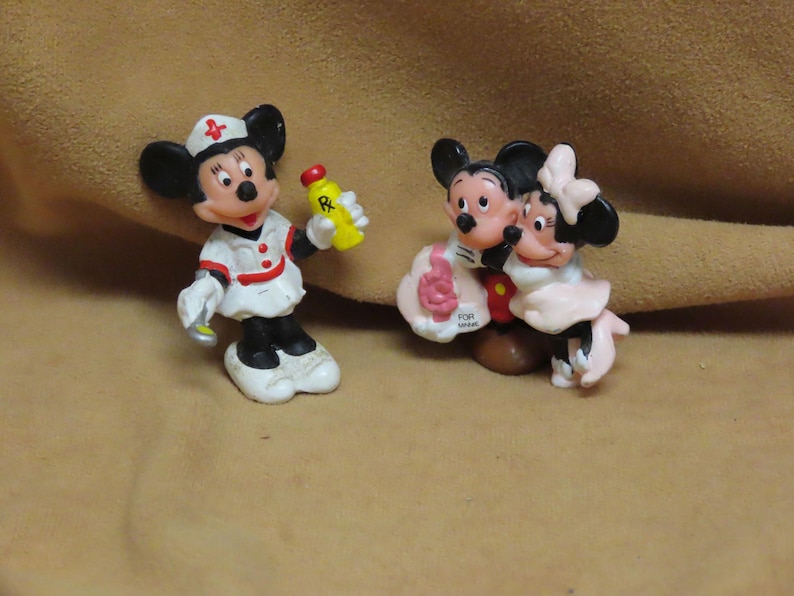 Puede incluir: Dos peque&ntilde;as figuras de Disney. Una es Mickey Mouse vestido de enfermera, sosteniendo una botella amarilla con una tapa roja. La otra figura es Mickey y Minnie Mouse abraz&aacute;ndose. Las figuras est&aacute;n sobre una superficie beige.