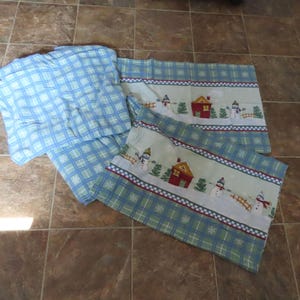 May include: A set of bedding items including a blue and white plaid patterned fabric piece and two rectangular pillowcases. The pillowcases feature a winter scene with snowmen, small houses, and green trees against a light background.