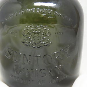 Puede incluir: Botella de vidrio verde oscuro de Suntory Whisky, Producto de Japón. La botella tiene texto en relieve y un escudo. El texto incluye "A blend of the choice whisky" y "1899". La botella es parcialmente visible.