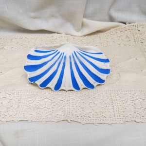 Puede incluir: Un plato blanco con forma de concha con rayas azules pintadas. El plato está sobre una superficie crema con bordes de encaje, evocando un tema costero o náutico.