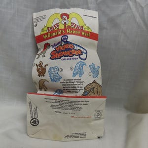 May include: A white paper bag with a McDonald's Happy Meal logo and a Disney Video Showcase Collection logo. The bag features a cartoon image of Ronald McDonald and a variety of animated characters from Disney's Dinosaur movie. The text on the bag reads "In computer-animated movies like Disney's "Dinosaur" characters are based on 3-D electronic models. Can you tell which model belongs to which character?"