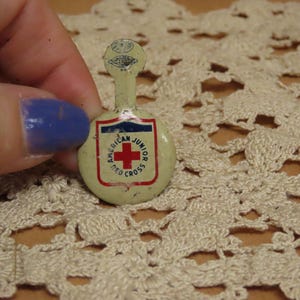 May include: A vintage cream-colored metal pin featuring a shield-shaped emblem with a red cross and the text American Junior Red Cross. The pin has a circular base with a small vertical tab at the top containing additional circular graphic details.