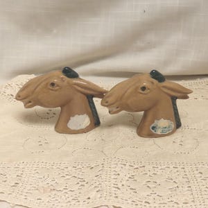 May include: A pair of ceramic horse head figurines. The figurines are tan with black accents on the mane and ears. One figurine has a white patch on the chest, and the other has a sticker. The horse heads are displayed on a lace tablecloth.