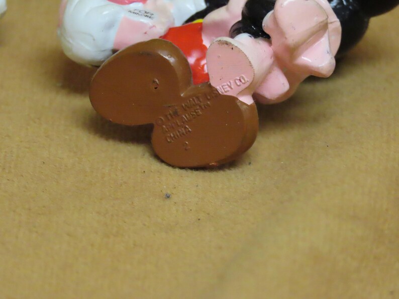 Puede incluir: Primer plano de la parte inferior de una figura de Mickey Mouse. Los pies de la figura son marrones, con el texto "&copy; THE WALT DISNEY CO. MADE IN CHINA" impreso en la parte inferior. Se ven los pies rosas y los zapatos negros de la figura.