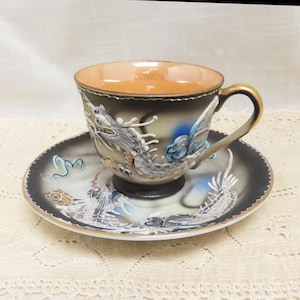 Op de afbeelding: Een vintage theekop en schotel set. De kop en schotel zijn zwart met een gedetailleerd drakenontwerp in zilver en blauw. De binnenkant van de kop is warm oranje en het handvat is goud.
