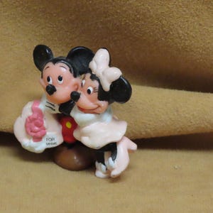 Puede incluir: Un juego de figuras con Mickey Mouse y Minnie Mouse en un abrazo amoroso. Mickey lleva una chaqueta negra y Minnie un vestido rosa. Una botella amarilla con tapa roja es sostenida por una mano enguantada. La base dice "FOR MINNIE".