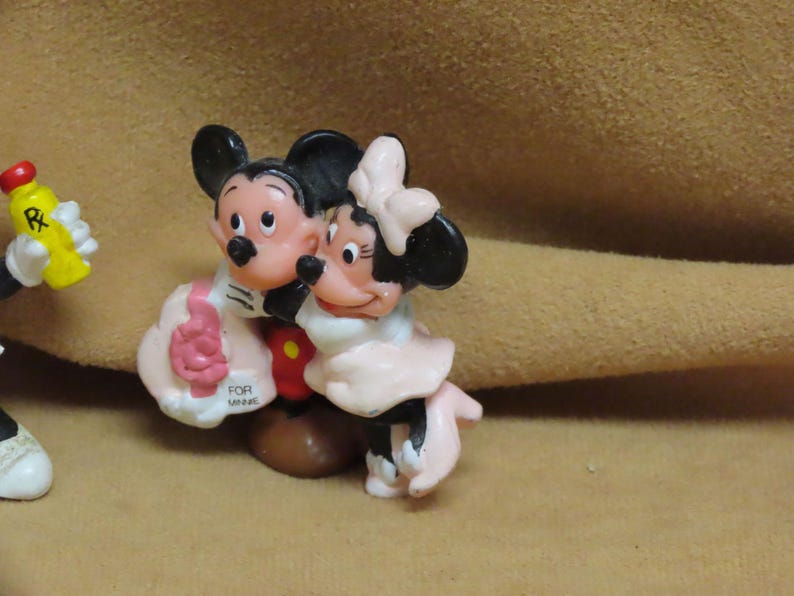 Puede incluir: Un juego de figuras con Mickey Mouse y Minnie Mouse en un abrazo amoroso. Mickey lleva una chaqueta negra y Minnie un vestido rosa. Una botella amarilla con tapa roja es sostenida por una mano enguantada. La base dice "FOR MINNIE".