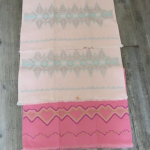 Op de afbeelding: Drie decoratieve textielstukken met gefranjerde randen. Twee zijn lichtroze met een herhalend patroon van turquoise, grijze en bruine geometrische vormen. De derde is felroze met een zigzagpatroon in goud, paars en roze.