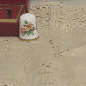 Puede incluir: Un dedal de porcelana blanca con un diseño floral en naranja, amarillo y verde, acentuado con una banda dorada. El dedal se exhibe junto a una caja de joyería roja sobre una superficie de encaje color crema.