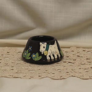 May include: A small, dark-colored ceramic bowl with a tapered shape. The exterior features a hand-painted white goat wearing a green collar with a yellow bell, surrounded by small blue flowers with green leaves painted on the black surface.