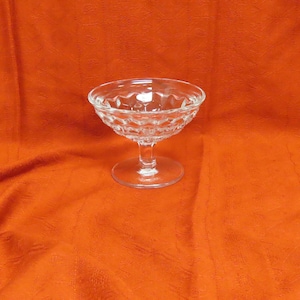 May include: A clear, stemmed glass with a diamond-cut pattern. The glass has a round bowl and a short stem, ideal for serving desserts or cocktails. The glass is set against an orange background.