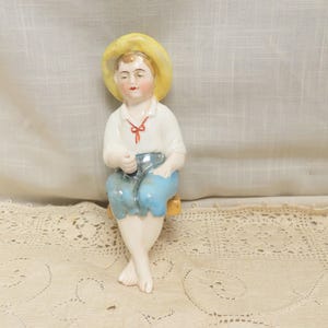 May include: A vintage porcelain figurine of a seated figure wearing a yellow hat, white shirt with a red bow, and blue pants. The figurine is resting on a small yellow seat. The figure is holding a small object.