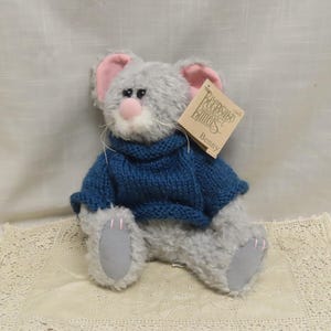 May include: A plush gray mouse toy wearing a blue knitted sweater. The mouse has pink ears, a pink nose, and gray paws. A tag that says "Keepsake Knitters Benny" is attached to the toy.