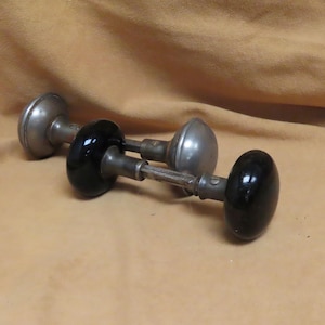 May include: Two antique door knobs. One is silver-toned metal with a rounded shape, and the other has a black, oval-shaped handle. The knobs are set against a tan background.