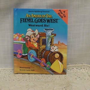 Peut inclure: Livre pop-up pour enfants intitulé "An American Tail Fievel Goes West". La couverture bleue présente un dessin animé de souris sur un train, avec un décor de désert et le texte "Westward Ho!". Illustré par Beverly Lazor-Bahr.
