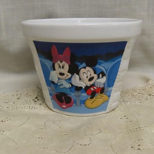 May include: White ceramic planter featuring a colorful image of Mickey Mouse and Minnie Mouse. The design shows the iconic characters in a blue and white setting. The planter is suitable for indoor or outdoor use.