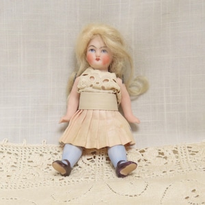 May include: A porcelain doll with blonde hair, blue eyes, and a painted pink face. The doll is wearing a white lace dress with a beige skirt and a beige belt. The doll is sitting on a white crocheted surface.