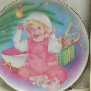 Könnte beinhalten: Dekorative Platte mit einem lächelnden Kind in rosa Latzhose und weißem Hut. Die Platte hat einen goldenen Rand und ein Weihnachtsthema, mit einem geschmückten Baum, einem verpackten Geschenk und einer rosa Schüssel.
