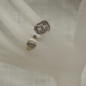May include: Two rings are displayed. One is silver-toned with a decorative design and small embellishments. The other ring is gold-toned with a textured surface. Both rings are on a white surface.