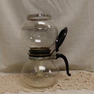 May include: A vintage glass coffee maker with a black handle and lid. The clear glass body has a bulbous bottom and a round top, with a decorative band around the middle. The coffee maker sits on a white surface.