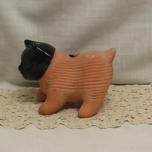 May include: A ceramic cat figurine with a black head and a peach-colored body. The cat is in a walking pose, with its body featuring horizontal ridges. The figurine is placed on a cream-colored lace doily.