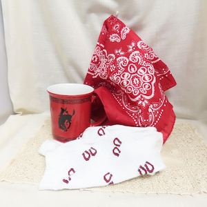 Puede incluir: Una taza de cerámica roja con la silueta negra de un caballo encabritado, acompañada de un pañuelo rojo y blanco con estampado de cachemira y un paño blanco decorado con un patrón repetitivo de pequeñas herraduras rojas en pares.