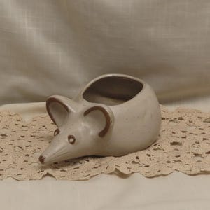 Puede incluir: Una maceta de cerámica de color beige claro con forma de ratón y acabado liso moteado. El ratón presenta dos orejas grandes y redondeadas delineadas en marrón, un hocico puntiagudo y ojos pequeños y sencillos. La parte superior es hueca.