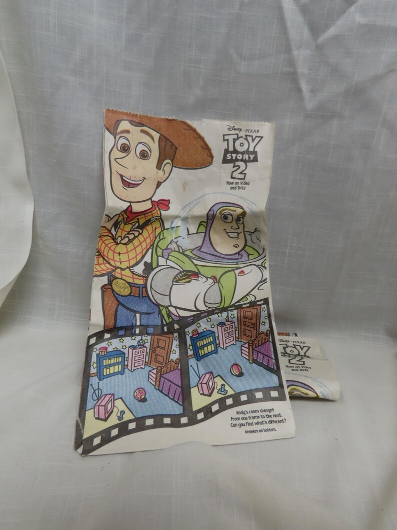 May include: A white paper bag with a fold-out design featuring Woody and Buzz Lightyear from the movie Toy Story 2. The bag has a spot-the-difference game with two images of Andy's room. The text on the bag reads "Disney Pixar Toy Story 2 New on Video and DVD".