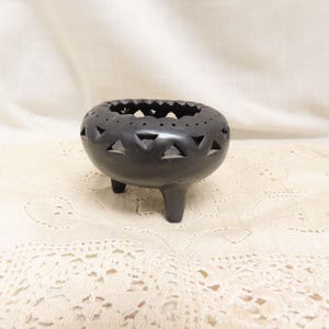 May include: A black ceramic incense burner with a round body and three short legs. The top edge has a scalloped design, and the sides feature triangular cutouts. The burner sits on a cream-colored, lace-patterned surface.