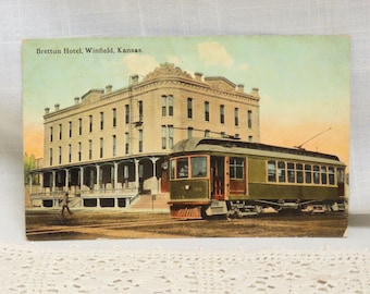 May include: A vintage postcard showing the Brettun Hotel in Winfield, Kansas. The hotel is a large, multi-storey building with a porch and a tram in front of it.
