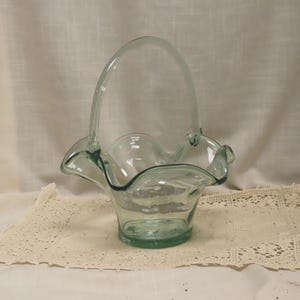 May include: A clear, light green glass basket with a handle. The basket has a ruffled edge and sits on a cream-colored lace doily. The handle arches gracefully above the basket, creating a decorative and functional piece.