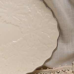 May include: A white ceramic plate with a scalloped edge and a leaf design. The plate has a silver rim.